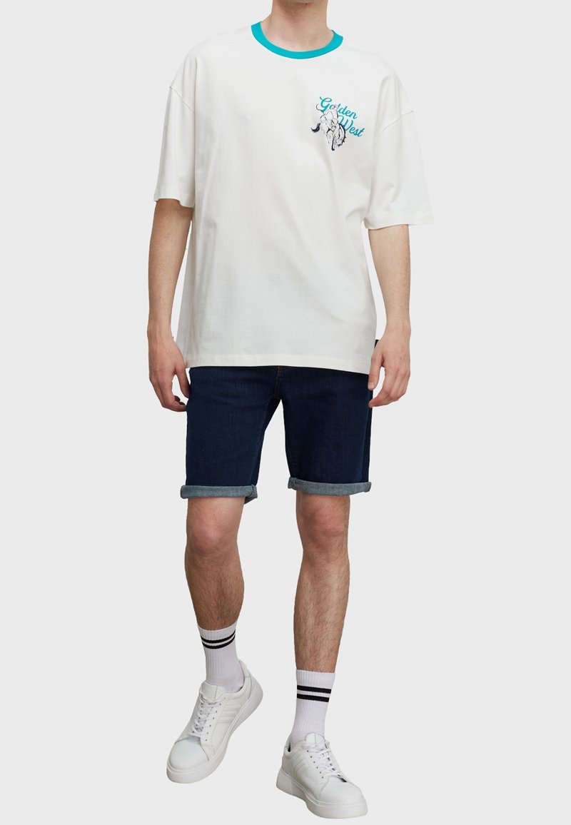 Hombre con una camiseta gráfica blanca de cuello aguamarina, pantalones cortos de mezclilla oscuros y doblados, calcetines blancos con rayas negras y zapatillas blancas.