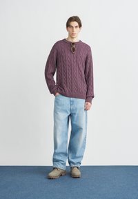 Μωβ πουλόβερ με πλεκτό σχέδιο, ανοιχτό μπλε φαρδιά τζιν, γκρίζες τσόκαρα. Το μοντέλο στέκεται σε μπλε πάτωμα μπροστά σε ανοιχτό φόντο. Γυαλιά ηλίου ακουμπισμένα στο πουλόβερ.