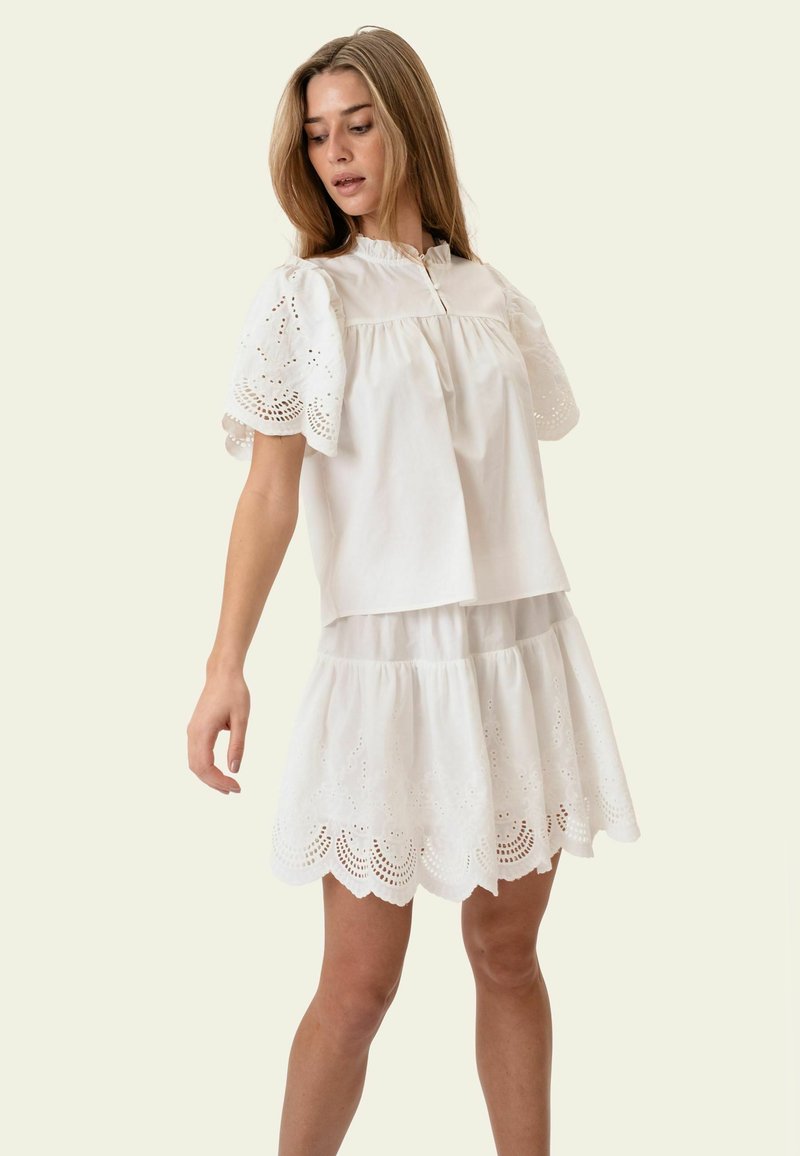 Femme portant un chemisier blanc à manches bouffantes en dentelle à œillets et une jupe à volants assortie, regardant vers le bas, debout devant un fond clair uni.