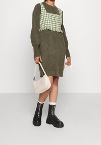 Ylimitoitettu oliivinvihreä neulemekko, jonka päällä vihreä-valkoinen mäyränhampaan kuvioitu lyhyt liivi; yhdistetty mustiin platform-saappaisiin ja tikattuun laukkuun.