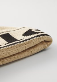 Strikket lue i beige med svart grafisk design. Den brettede kanten avdekker et teksturert strikkemønster som viser et mykt og varmt materiale.