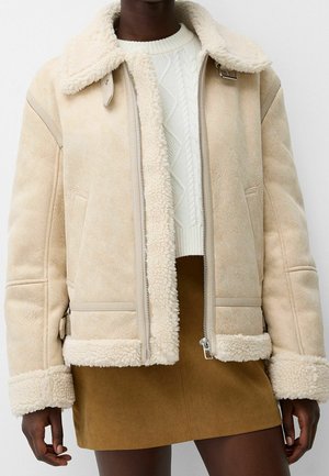 Giacca beige in shearling con collo e polsini morbidi, indossata sopra un maglione bianco a trecce e una gonna in suede marrone.
