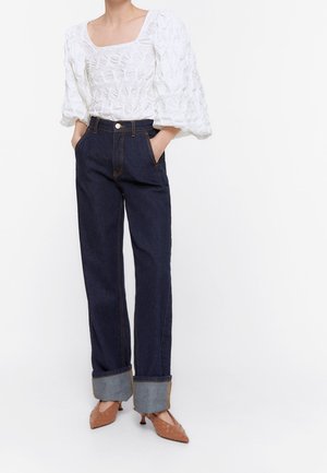 Weiße Bluse aus gekräuseltem Stoff mit quadratischem Ausschnitt und puffigen Ärmeln, kombiniert mit dunkelblauen Jeans mit geradem Bein, Manschetten und Taschen.