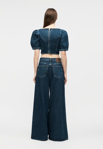 Γυναίκα με μακριά μαλλιά που φοράει σκούρο μπλε τζιν cropped τοπ με φουσκωτά μανίκια και τζιν παντελόνι ψηλόμεσο με φαρδιά πατ από πίσω.