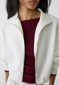 Chaqueta bomber blanca con cremallera y cuello simulado, combinada con una camiseta ajustada burdeos. Tejido suave con un corte relajado y acentos de canalé.