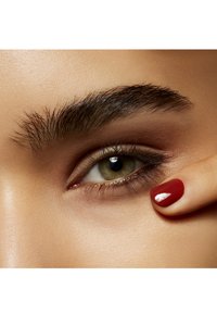 Close-up van een groen oog met gedefinieerde wimpers, zachte bruine oogschaduw en een verzorgde hand met rode nagellak die het onderste ooglid aanraakt.