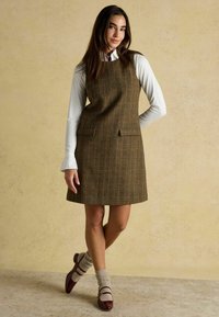 Braun karierte A-Linien-Kleid mit seitlichen Taschen, über einem weißen Langarmshirt getragen. Das Model kombiniert es mit burgunderroten Schuhen und grauen Socken.