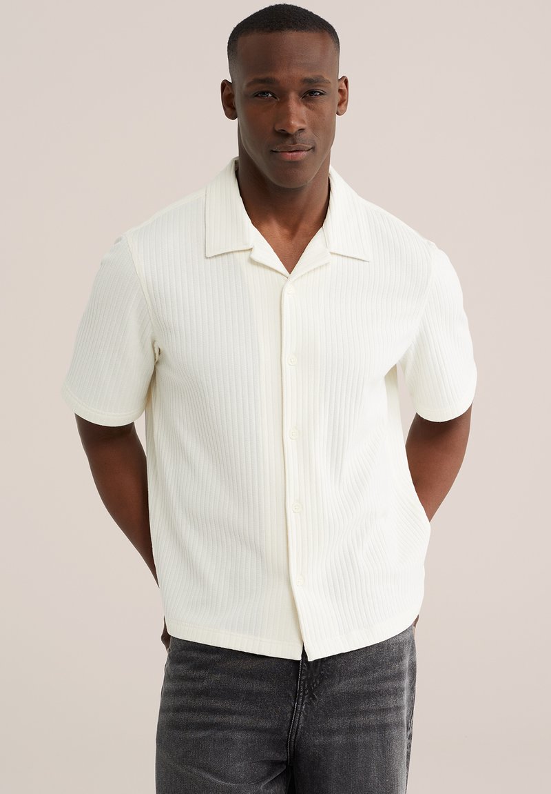 Homme portant une chemise blanche côtelée à manches courtes boutonnée et un jean foncé, debout les mains dans le dos sur un fond uni.