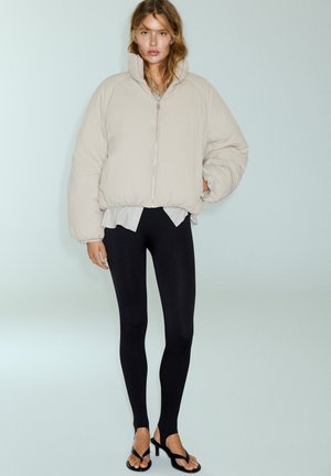 Femme portant une doudoune beige, des leggings noirs avec étriers, des sandales noires à talons et une chemise rayée en dessous, debout devant un fond uni.