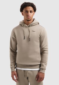 Jonge man met krullend haar, gekleed in een beige hoodie en bijpassende broek, staande tegen een eentonige lichtgrijze achtergrond.