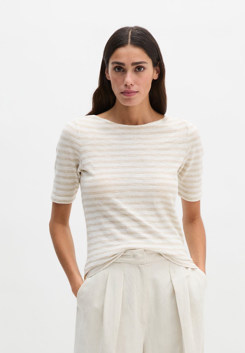 Kobieta o długich ciemnych włosach, ubrana w beżowo-biały pasiasty top z krótkim rękawem i jasnobeżowe plisowane spodnie, stojąca z rękami w kieszeniach.