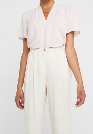 Femme portant un chemisier rose pâle à manches courtes et col en V, rentré dans un pantalon blanc plissé, ample et taille haute.