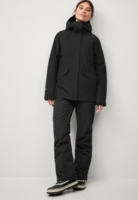 Black waterproof jacket with a high collar, two front pockets, and a cinched hood, paired with black pants and green-accented boots.