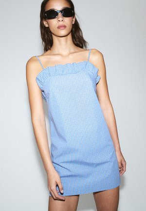 Femme portant des lunettes de soleil noires et une mini-robe bleue à carreaux sans manches avec un col à volants, se tenant contre un mur uni.
