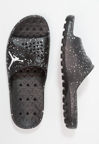 Mustat liukusandaalit valkoisen maalaustahra-muotoilun, teksturoidun pohjan ja hihnassa olevan valkoisen Jumpman-logon kanssa, esiteltynä ylhäältä ja sivulta.