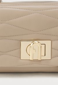 Bēša ādas soma ar viļņotu kviltētu tekstūru, zelta toņa aizdare ar logotipa detaļu. Gluda virsma un kārtīgas šuves.