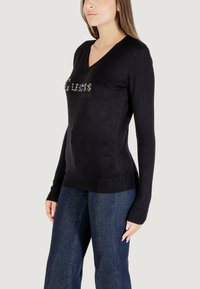Schwarzer V-Ausschnitt-Pullover mit langen Ärmeln und einem Strass-Logo "GUESS". Das Material ist glatt, mit einem gerippten Bund und Bündchen. Kombiniert mit Jeans.