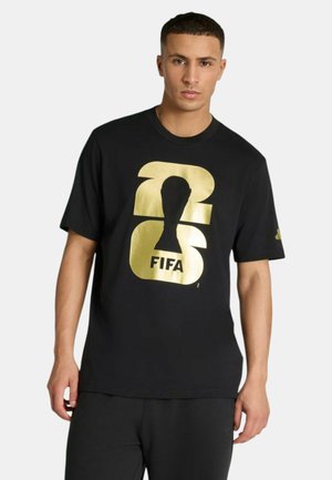 Uomo con maglietta nera con grande numero "23" dorato e silhouette del trofeo FIFA stampate sul davanti, in posa su sfondo bianco.