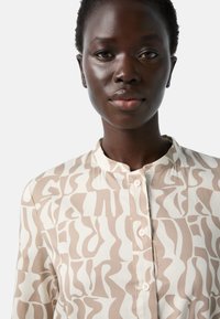 Langärmlige Bluse in Creme mit einem abstrakten beigen Muster, ausgestattet mit einem Stehkragen und einer vorderen Knopfleiste. Weicher Stofftextur.