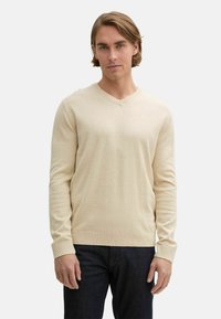 Beige V-Ausschnitt Pullover mit langen Ärmeln, aus einem weichen Stoff hergestellt. Verfügt über einen gerippten Saum und Ärmelsäume, entworfen für eine figurschmeichelnde Passform.