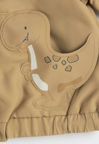 Tissu beige avec une applique de dinosaure en ton beige, ornée de détails en fil bleu et d'accents blancs. Le design inclut une tête arrondie et un contour texturé.