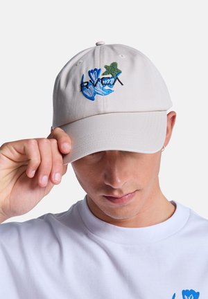 Persoon met een witte pet met blauwe en groene bloemenborduursels, die de rand rechtzet, gekleed in een wit shirt met een blauw bloemenpatroon.