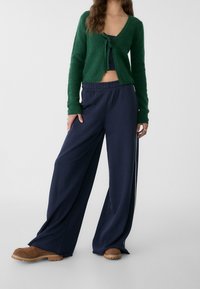 Cardigan verde peloso con chiusura a cravatta abbinato a pantaloni larghi blu navy. Scarpe marroni con suola texturizzata completano l'outfit.