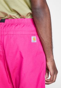 Person som bär ljusrosa Carhartt-shorts och ljusgrön skjorta, med högerarmen avslappnat vid sin sida mot en vit bakgrund.