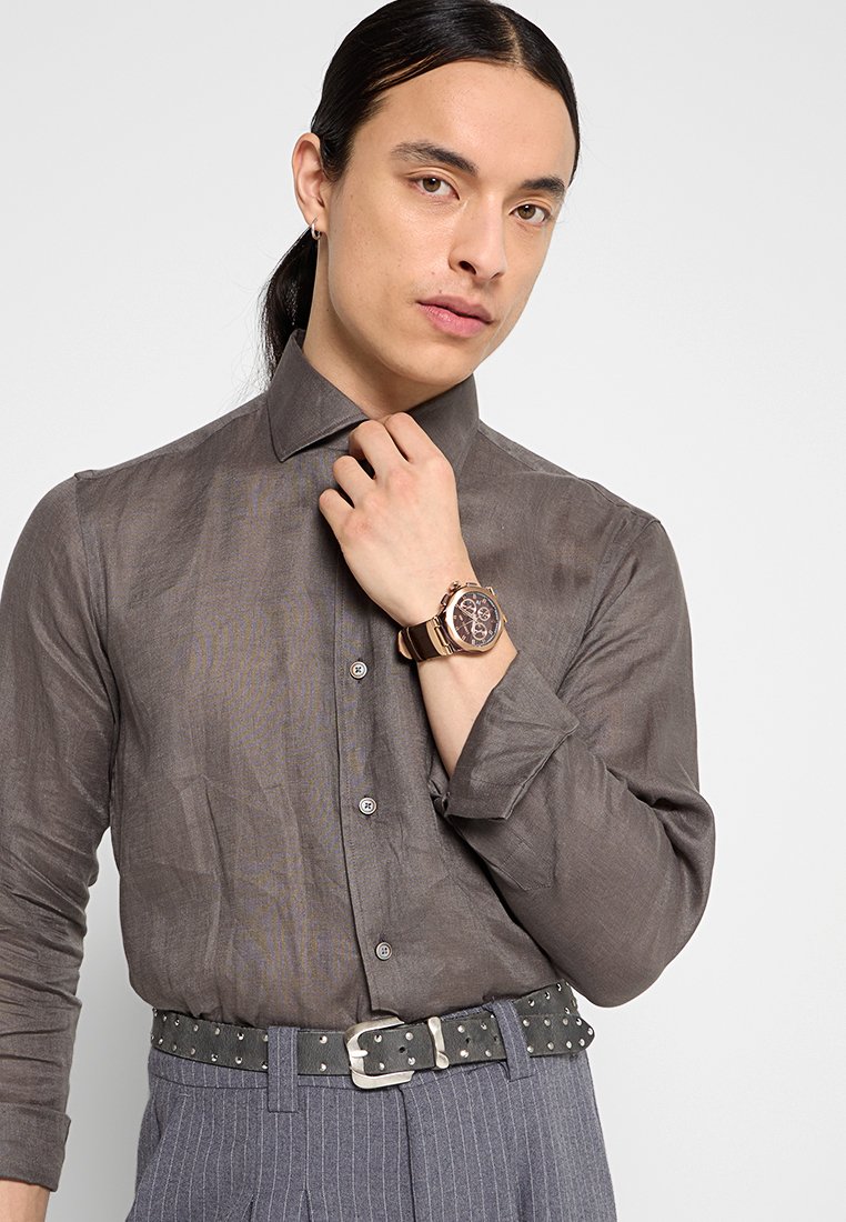 Jeune personne avec de longs cheveux noirs portant une chemise grise à boutons, ajustant son col, avec une montre en rose gold et une ceinture noire cloutée, ainsi qu'un pantalon à rayures.