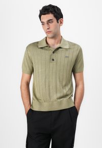 Olivgrön ribbad polo med treknappsstängning, korta ärmar och Lacoste-logotyp på vänster bröst, kombinerad med svarta byxor.