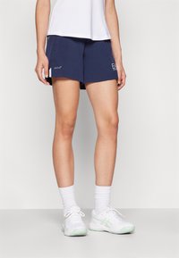 Calções de atletismo azul-marinho com bolsos laterais, apresentando um design elegante e detalhe do logótipo. Combinados com sapatilhas brancas e meias brancas caneladas.