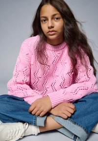 Rosa strikkegenser med et teksturert, åpent maskerdesign og ribbestrikkede mansjetter, kombinert med blå denimjeans og hvite joggesko.