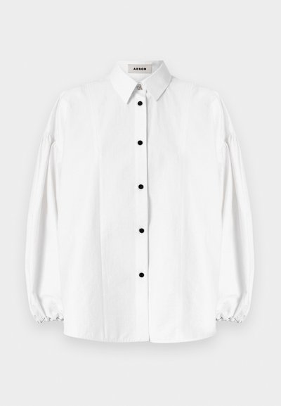 Camisa blanca de botones con mangas largas y abullonadas. Presenta botones negros, un cuello clásico y costuras verticales sutiles. Textura de tela suave.