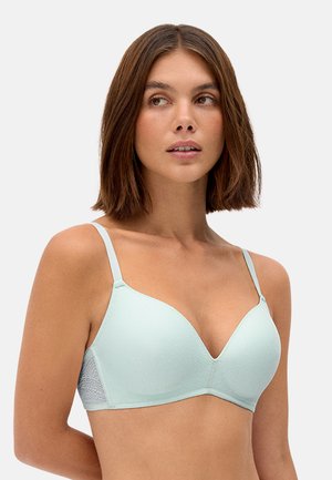 Mujer con cabello castaño hasta los hombros, que lleva un sujetador acolchado azul claro con tirantes ajustables, sobre un fondo liso.