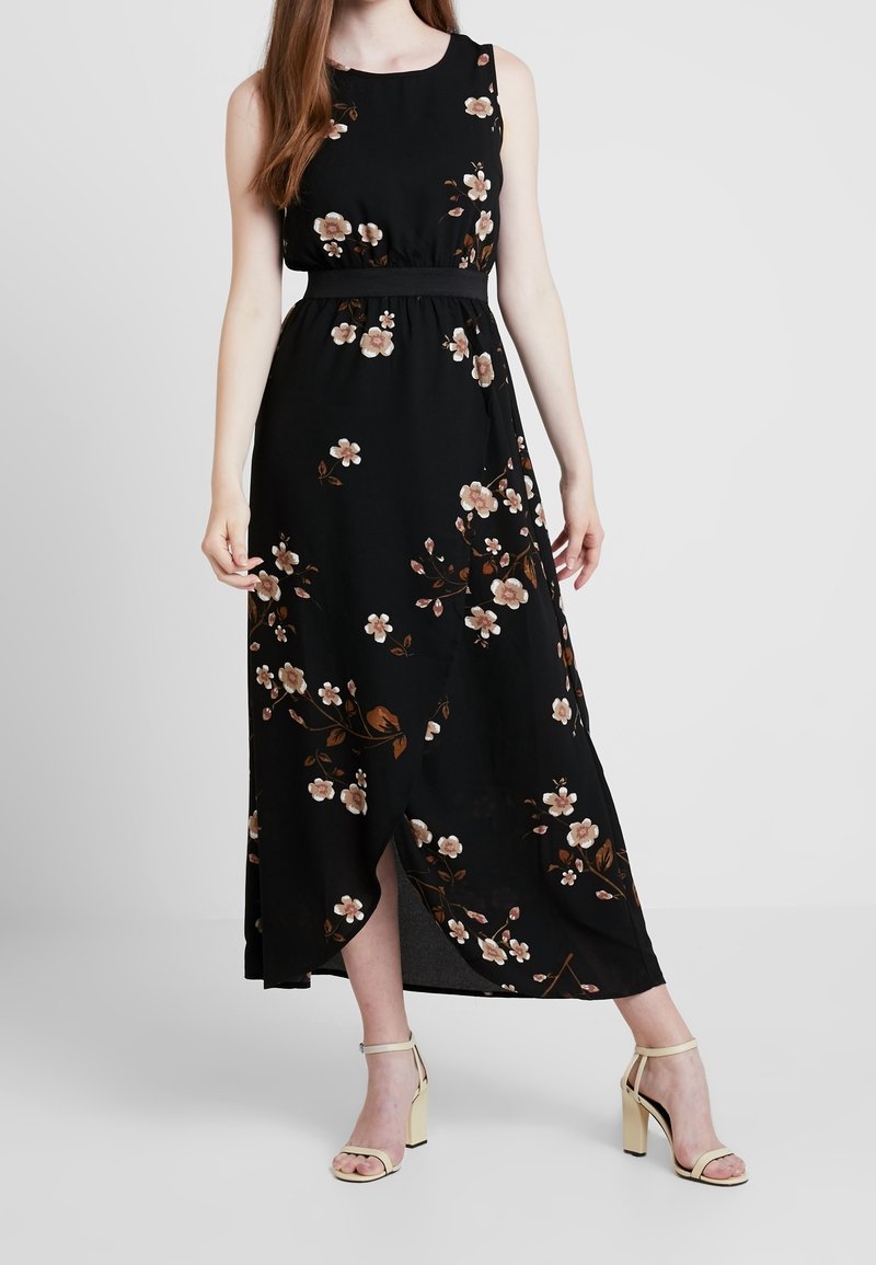 Nainen, joka on pukeutunut ilman hihoja mustaan maxi-mekkoon, jossa on beige kukkakuosi ja musta vyötärövyö, yhdistettynä beigeihin korkokenkiin.