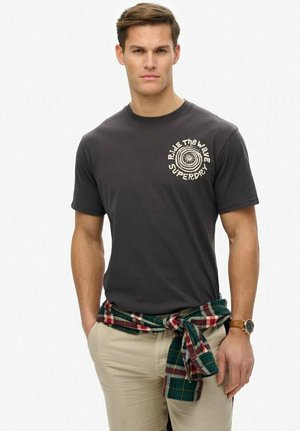 Gray short-sleeve t-shirt with white graphic on the chest, paired with beige trousers and a green and red plaid shirt tied around the waist.
