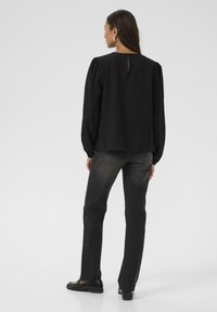 Blouse noire texturée avec des manches bouffantes et un détail en clé à l'arrière, associée à un jean foncé et des chaussures noires brillantes.