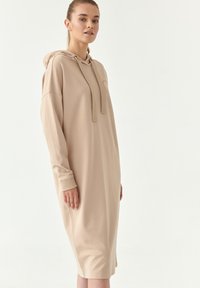 Abbigliamento a felpa beige con maniche lunghe, una tasca a marsupio e cordino regolabile. Tessuto morbido con una vestibilità rilassata, che arriva a metà polpaccio.