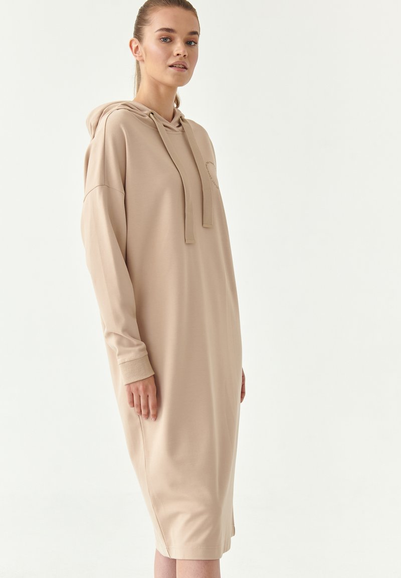 Abbigliamento a felpa beige con maniche lunghe, una tasca a marsupio e cordino regolabile. Tessuto morbido con una vestibilità rilassata, che arriva a metà polpaccio.