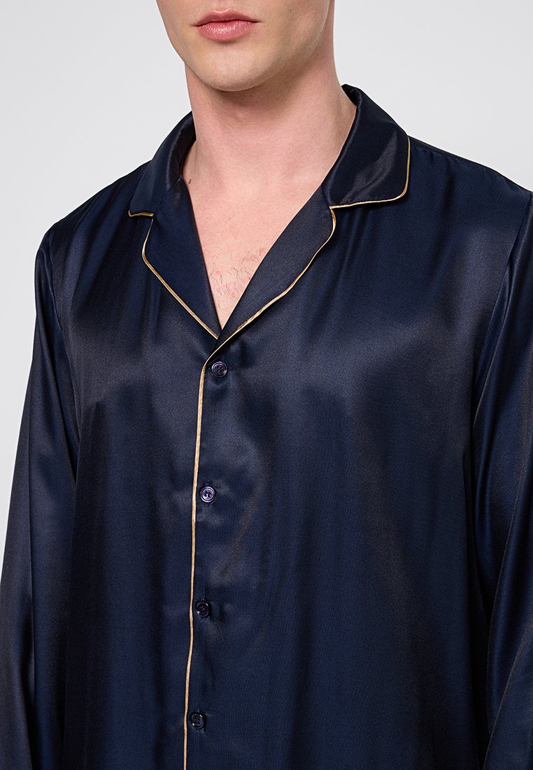 Homme portant un haut de pyjama en satin bleu marine avec des bordures et des boutons dorés, montré de la nuque au milieu du torse sur un fond uni.