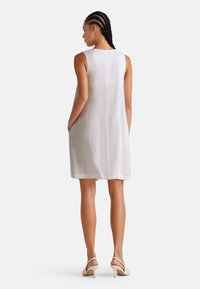 Weißes Leinenkleid ohne Ärmel mit lockerer Passform, das über Seitentaschen und eine subtile Textur verfügt. Kombiniert mit cremefarbenen Absatzsandalen.