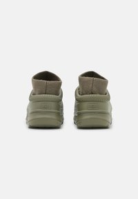 Gröna slip-on skor med en slitstark gummisula och en mjuk, texturerad krage i tyg. UGG-logotypen är präglad på baksidan.