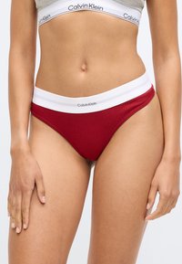 Punane ribatatud puuvillane string, millel on valge elastne vöökoht, millel on "Calvin Klein" logo. Sile tekstuur, madala tõusuga disain.