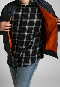 Giacca nera con rivestimento arancione, apertura con zip. Indossata sopra una camicia a quadri in nero, bianco e grigio. Fit casual con dettagli di cucitura visibili.