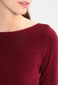 Top in magenta a maglia con scollatura ampia a costine e texture delicata. Maniche leggermente aderenti e corpo liscio. Nessun motivo o abbellimento visibile.