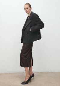 Abrigo de lana de doble botonadura en gris oscuro, combinado con una falda de cuero marrón ajustada con una abertura lateral, complementado con zapatos de tacón alto negros.