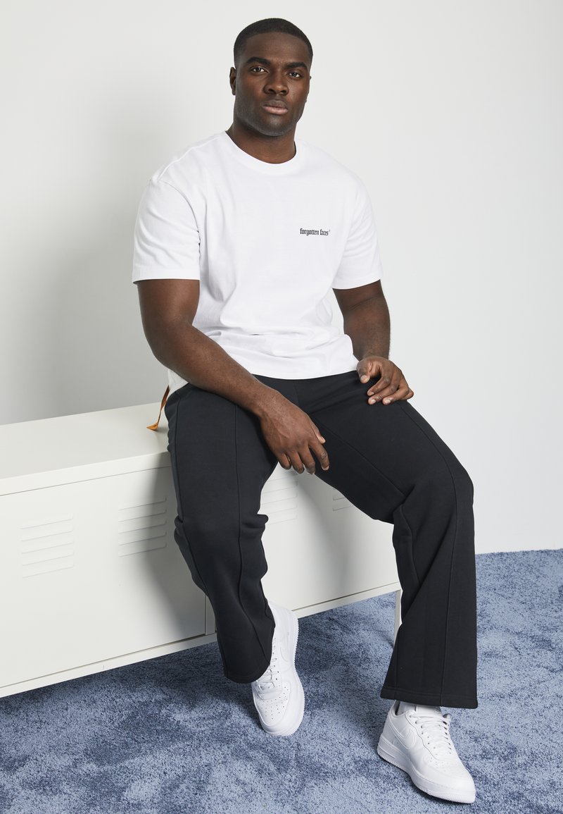 White cotton T-shirt with black logo print, paired with black pants and white sneakers. Smooth texture and classic fit, seated on a white surface.