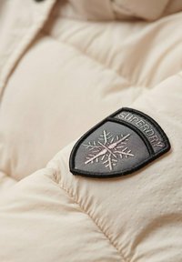 Beżowa kurtka puchowa o pikowanej teksturze. Posiada czarno-szary naszywkę z wzorem płatka śniegu i napisem "SUPERDRY".