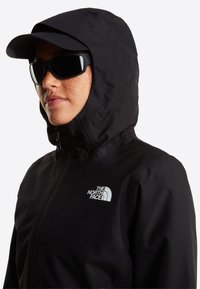 Άτομο που φοράει μαύρο μπουφάν North Face με κουκούλα, μαύρο καπέλο και μαύρα γυαλιά ηλίου, στραμμένο προς τα αριστερά σε λευκό φόντο.