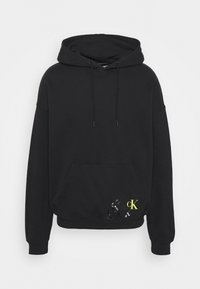Sudadera con capucha negra, con un bolsillo frontal, capucha con cordón y un llamativo detalle en logo amarillo y negro. Tejido suave, corte relajado.